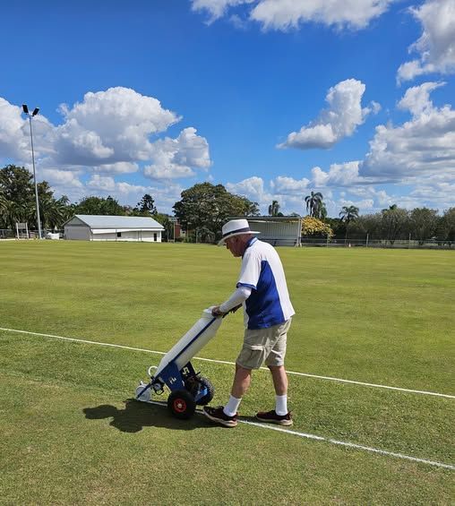 A man in a white hat and blue shirt pushes a manual lawn roller across a striped croquet court under a partly cloudy sky