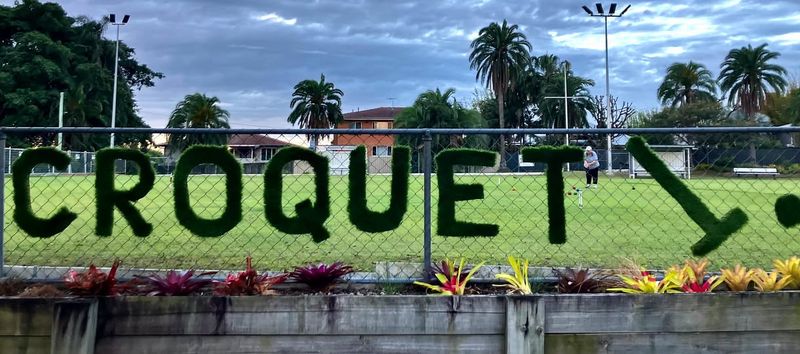 Green topiary letters spelling "CROQUET" on a chain-link fence, with a player and croquet hoops on a lawn behind it